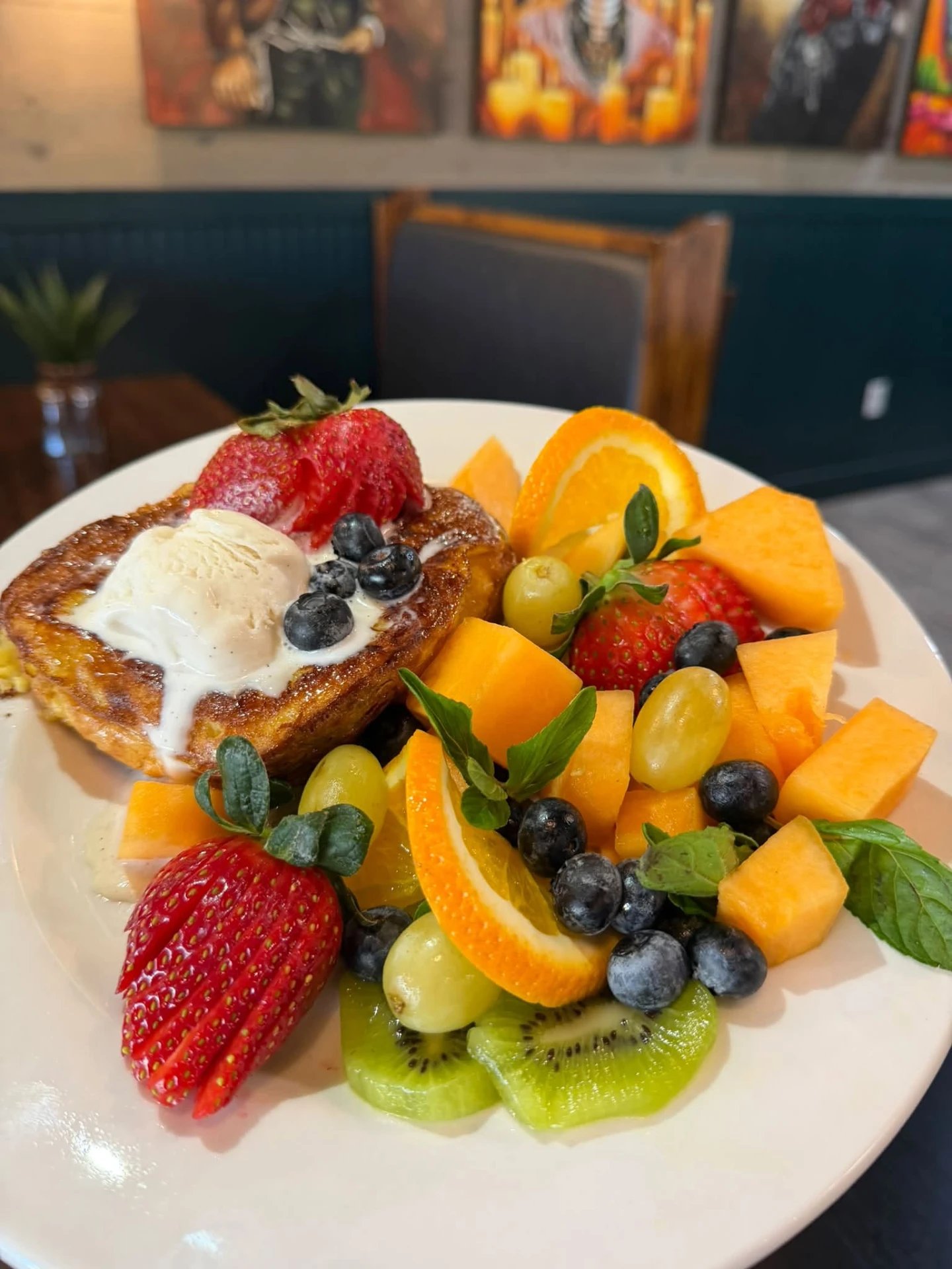 Golden French toast topped with melting vanilla ice cream, blueberries and a strawberry, beside a plate of fresh seasonal fruit
