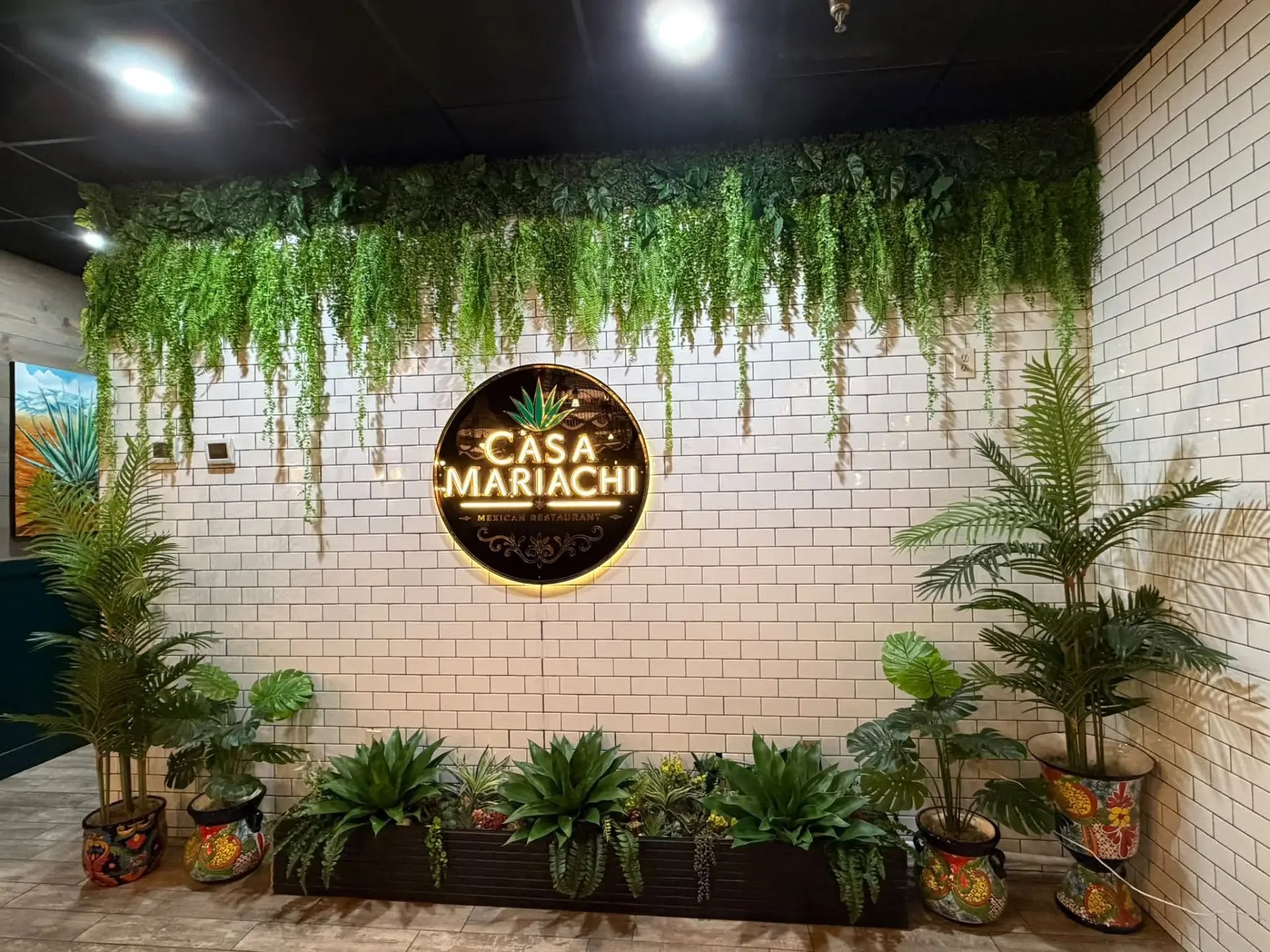 The Casa Mariachi dining room: a lit Casa Mariachi sign on a white tiled wall, framed by greenery and painted ceramic planters